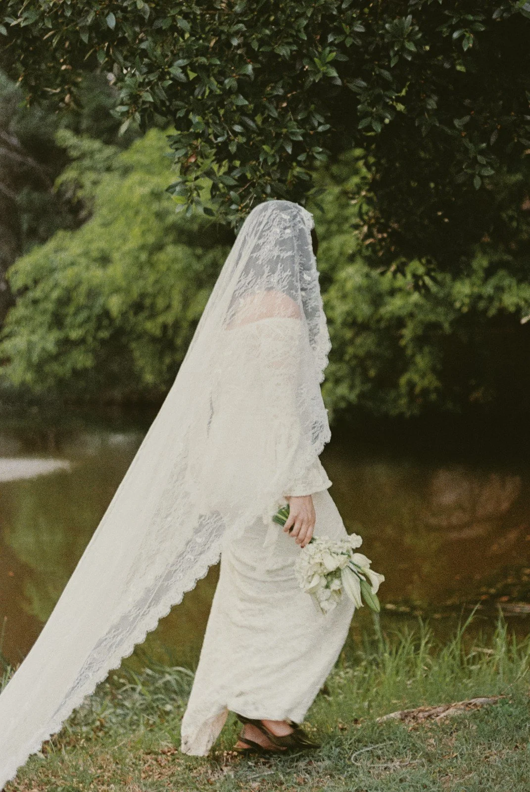 A bride wearing a white lace wedding dress and veil, holding a bouquet of white flowers, standing outdoors near a body of water and surrounded by green trees.
