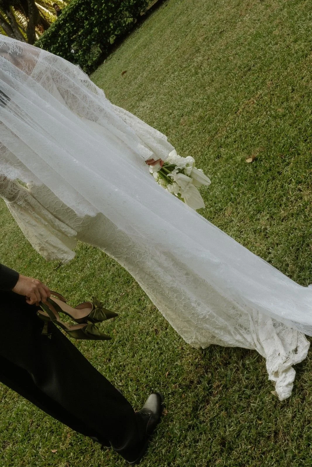 Wedding dress with lace detail, beige high heels with bows, and a bouquet of white flowers on the grass outdoors.