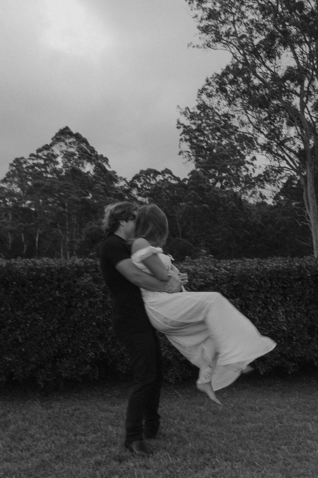 A couple kissing outdoors at sunset, with the woman wearing a dress and being lifted by the man, against a background of trees and cloudy sky.