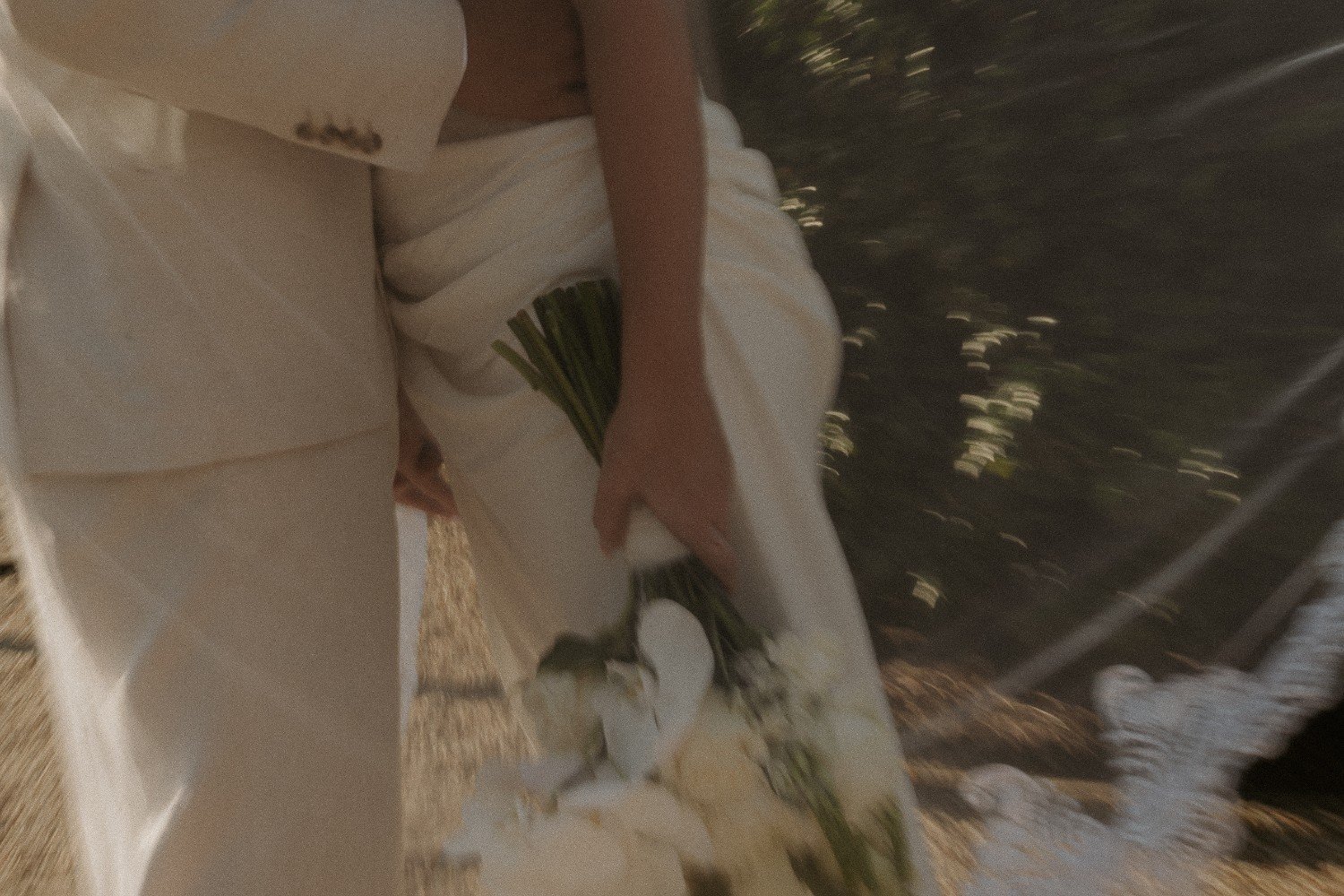A bride wearing white wedding gown holding a bouquet of white roses, outside near trees and sunlight.