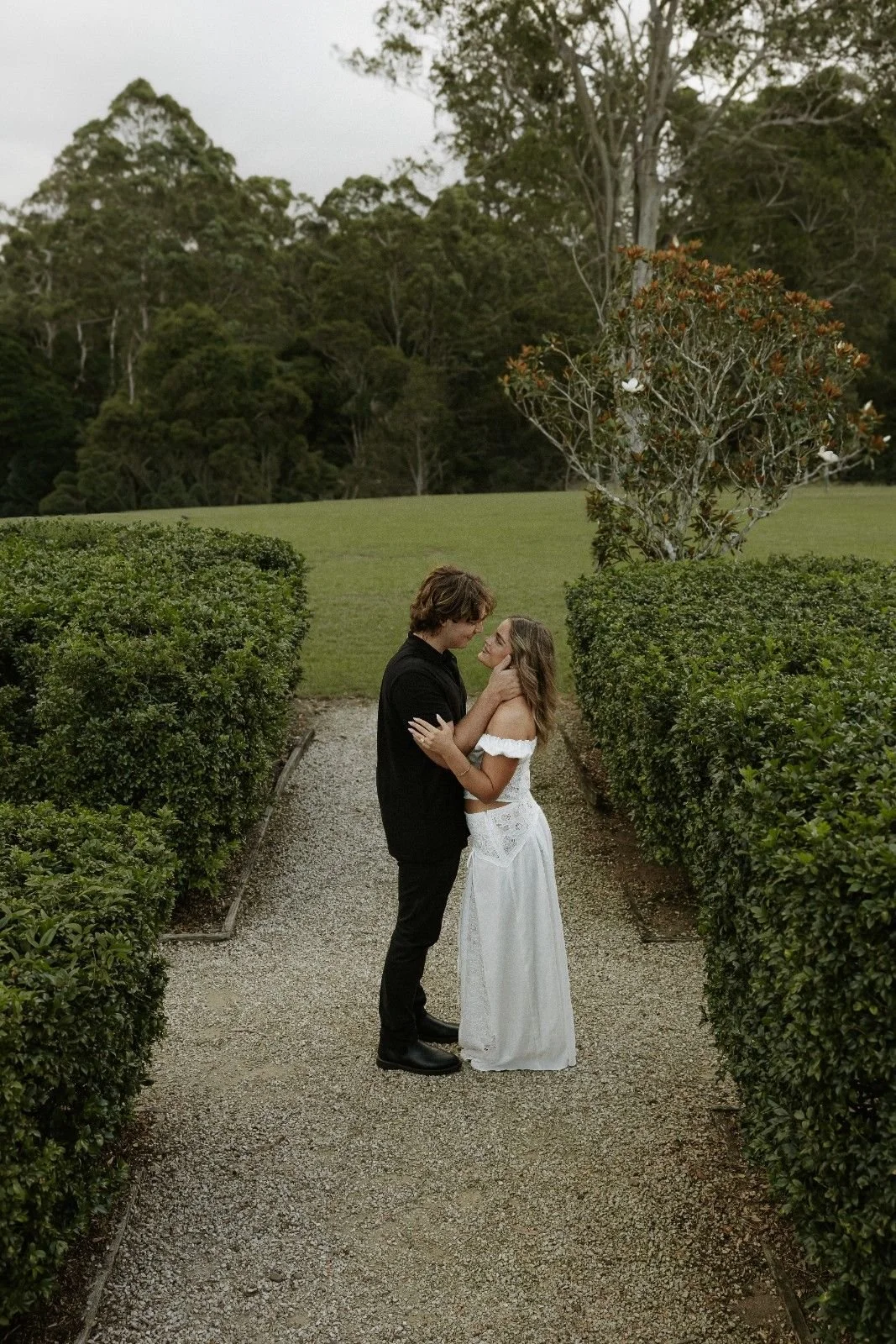 A couple, dressed in wedding attire, stands close together in a garden pathway surrounded by green bushes and trees, gazing into each other's eyes.