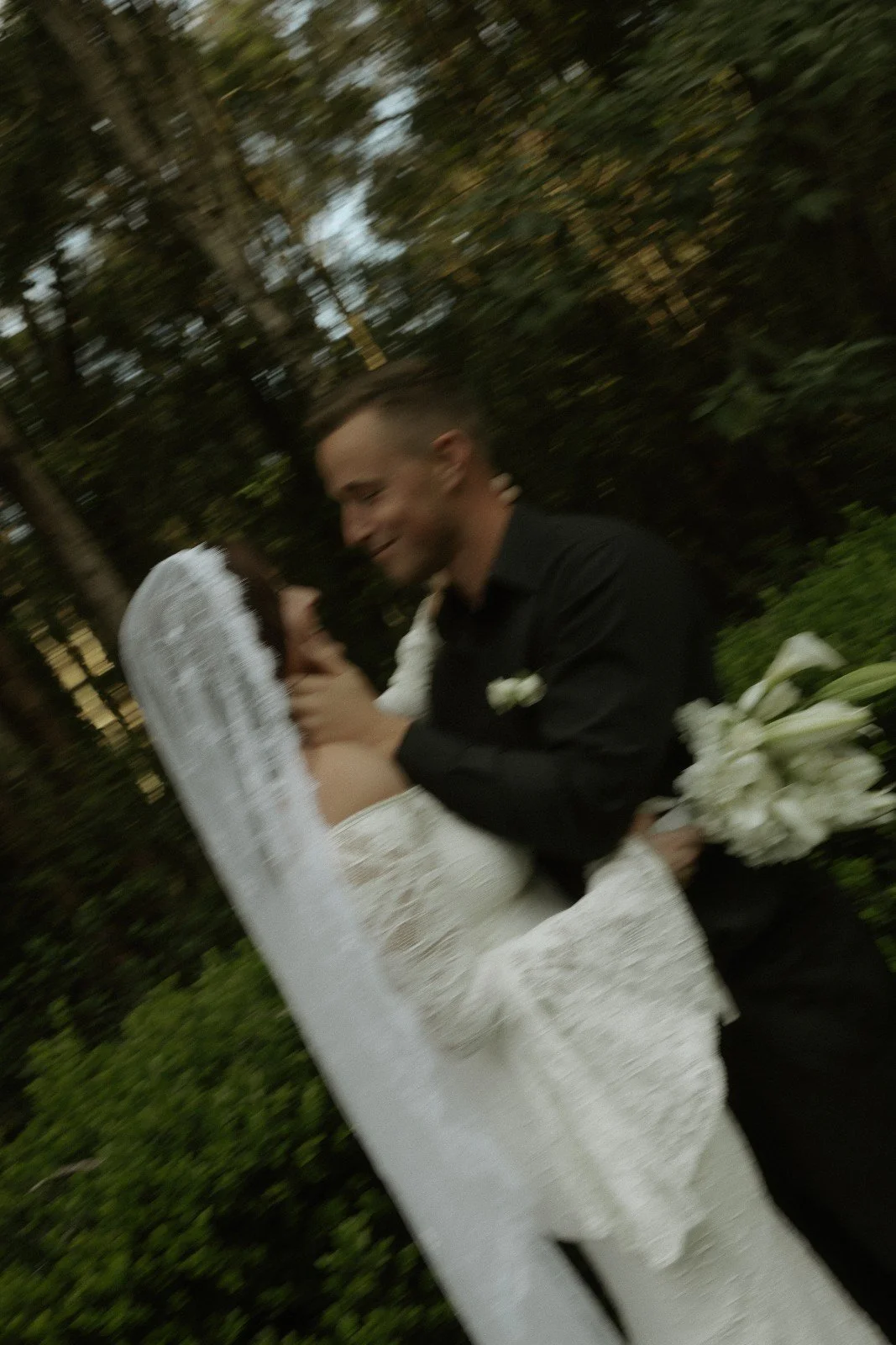 A bride and groom are dancing outdoors, smiling and holding each other, with trees and greenery in the background.