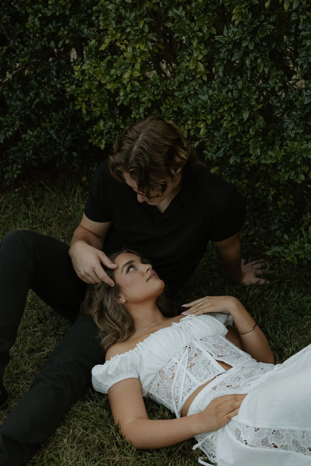 A man and a woman are lying on the grass in front of bushes. The woman is looking up at the man, who is touching her forehead gently. The woman is wearing a white lace dress, and the man is wearing a black shirt.