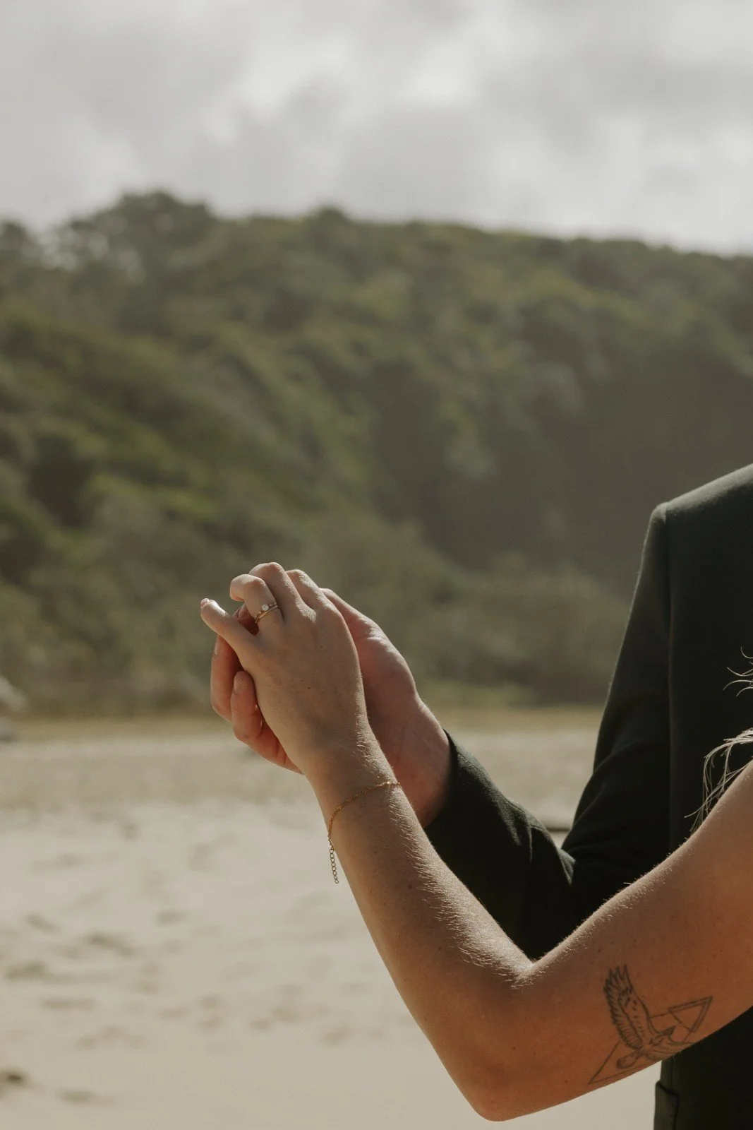 A couple holding hands outdoors, with a blurred background of a beach and green hills under an overcast sky.