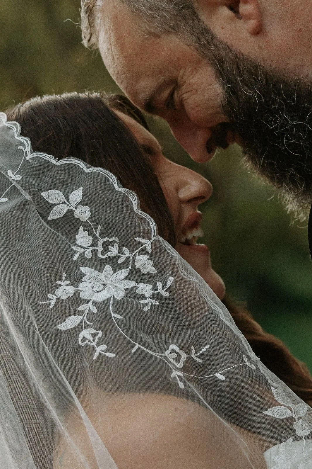 A bride and groom close together, sharing a moment, with the bride wearing a sheer, embroidered veil and the groom with a beard.