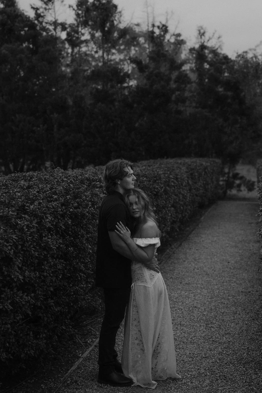 A black and white photo of a young couple standing on a gravel path, embracing each other, with trees and bushes in the background.