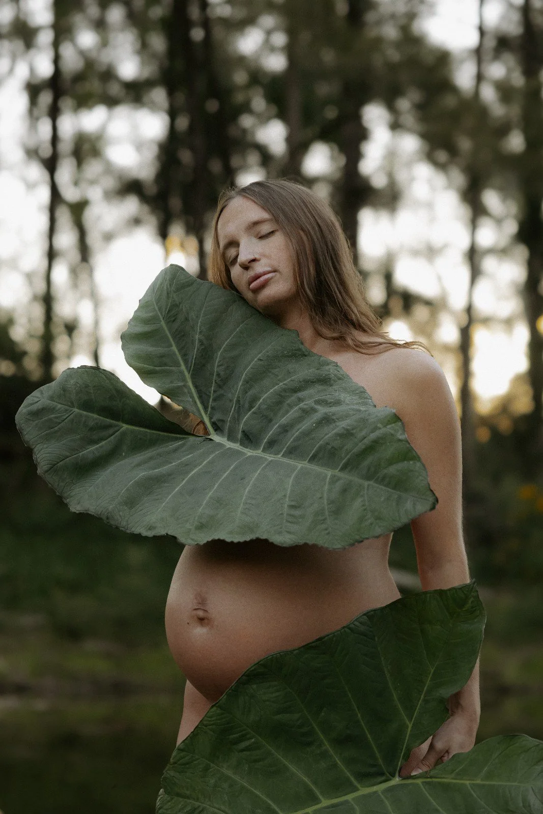 A woman with long brown hair is standing outdoors among large green leaves in a forest, with her eyes closed and her face resting against a leaf.