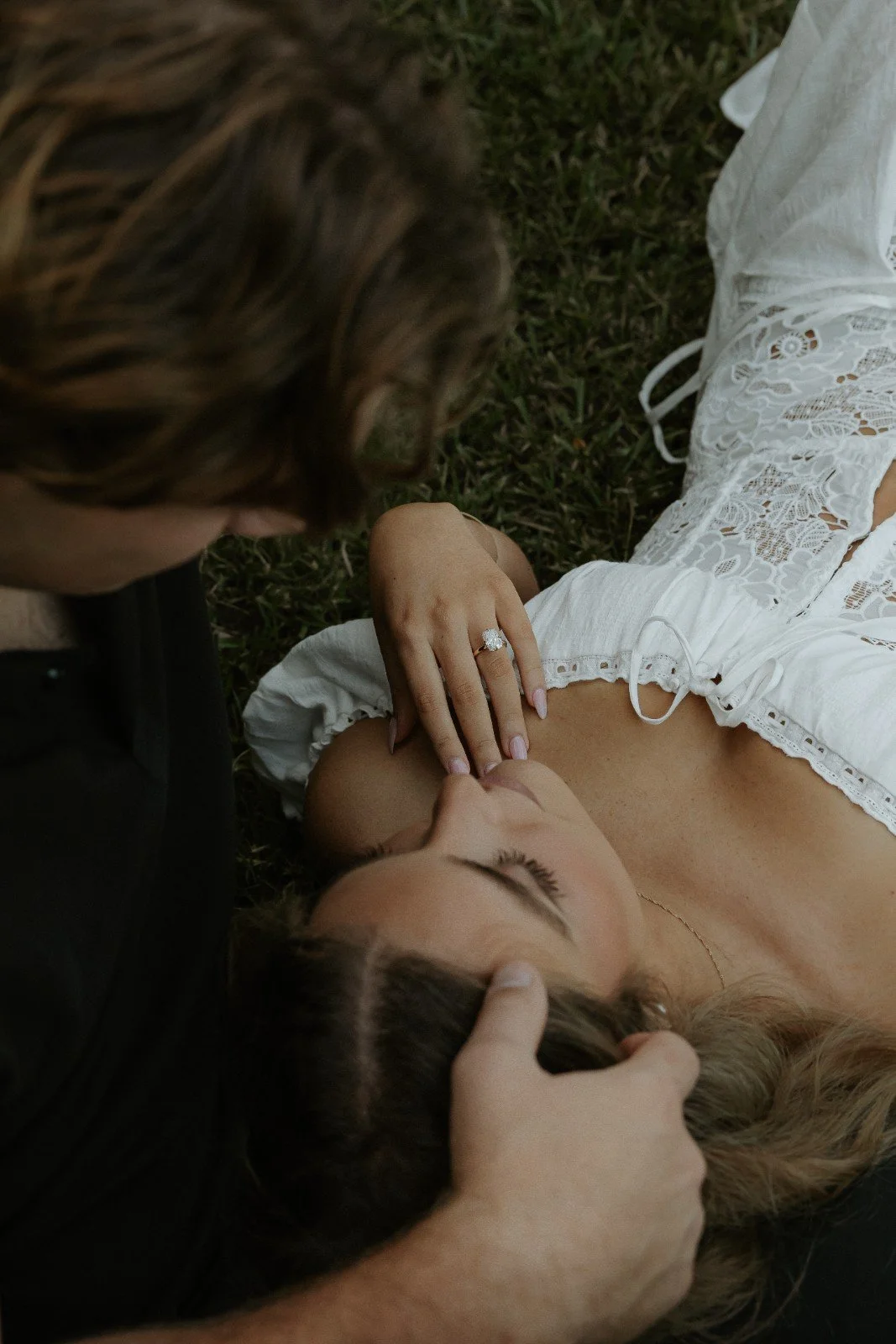 Two women laying on grass, one touching the other's face and the other wearing a wedding ring, with a close-up of their faces and one woman's hand on her chest.
