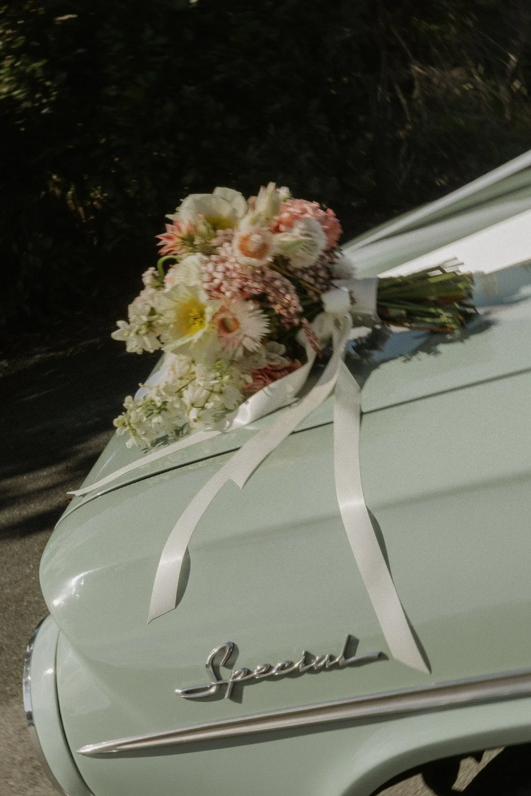 A vintage mint green car with a bouquet of white and pink flowers attached to the hood in front of a black shrub background.