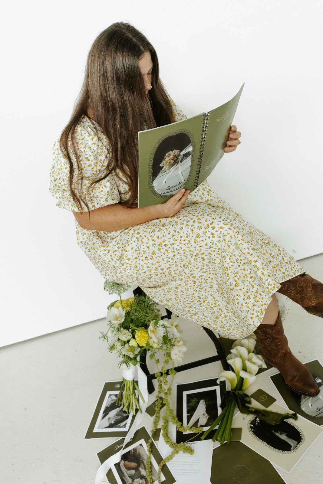 Robyn Small in a floral dress sitting on a black chair, reading a large hardcover book, with flowers, photographs, and papers arranged on the floor around her.