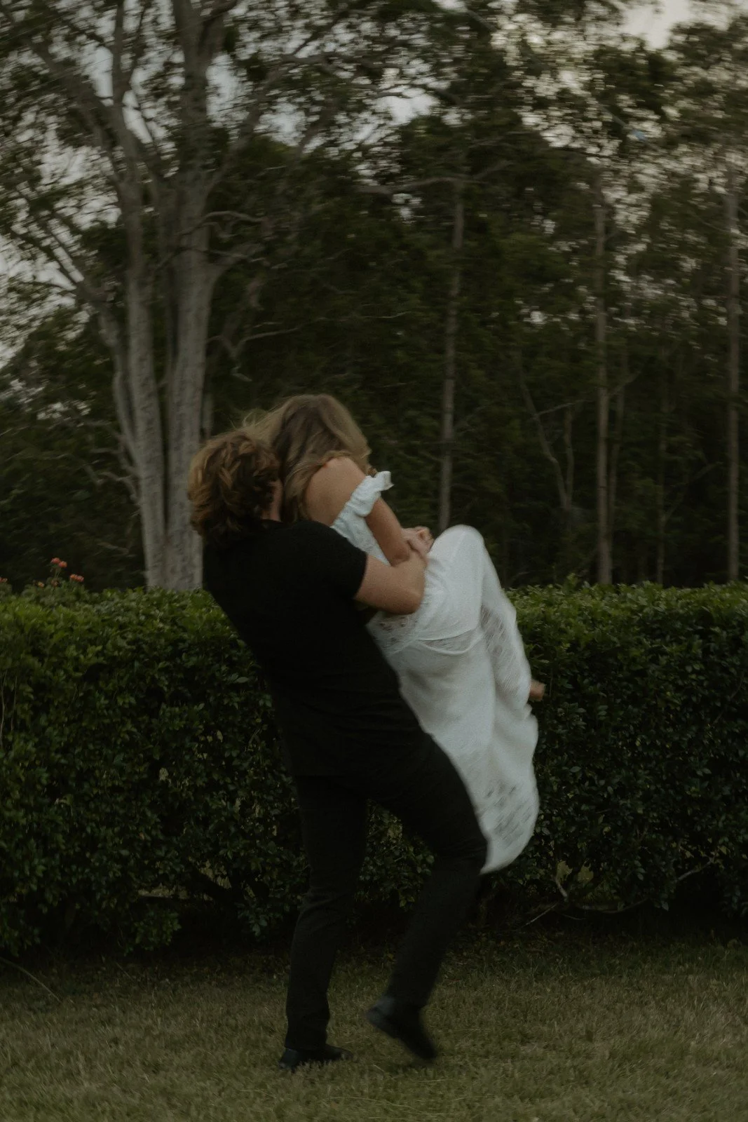 Person in black shirt lifting a woman in a white dress outdoors near bushes and trees.