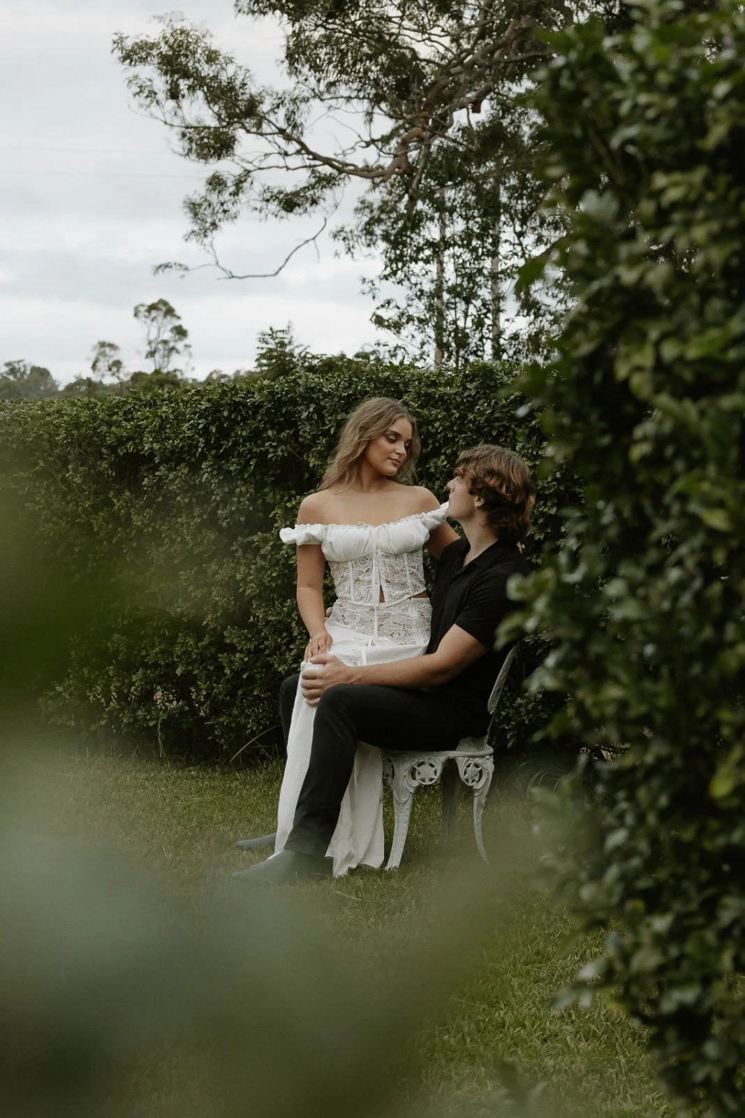 A woman in a white lace dress standing and a man in a black shirt sitting on a vintage chair outdoors, surrounded by greenery and bushes, with trees in the background.