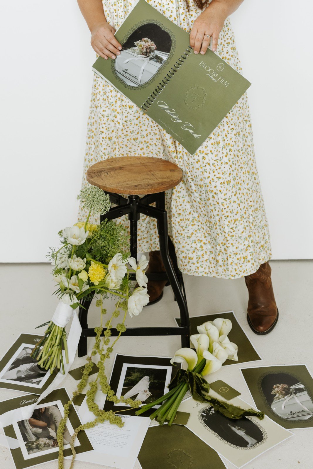 Woman in a white dress with yellow floral pattern holding a photography album titled 'Votivare Tuas' from Bloom Film with a bouquet of white flowers on a black stand, surrounded by flower images and photographs on the floor.