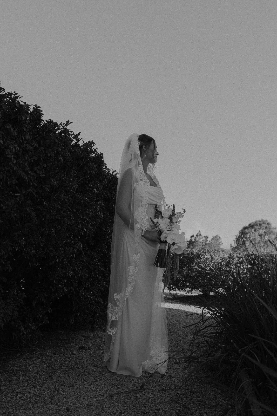 A bride in a wedding dress holding a bouquet, standing outdoors near bushes and trees, with a clear sky in the background.