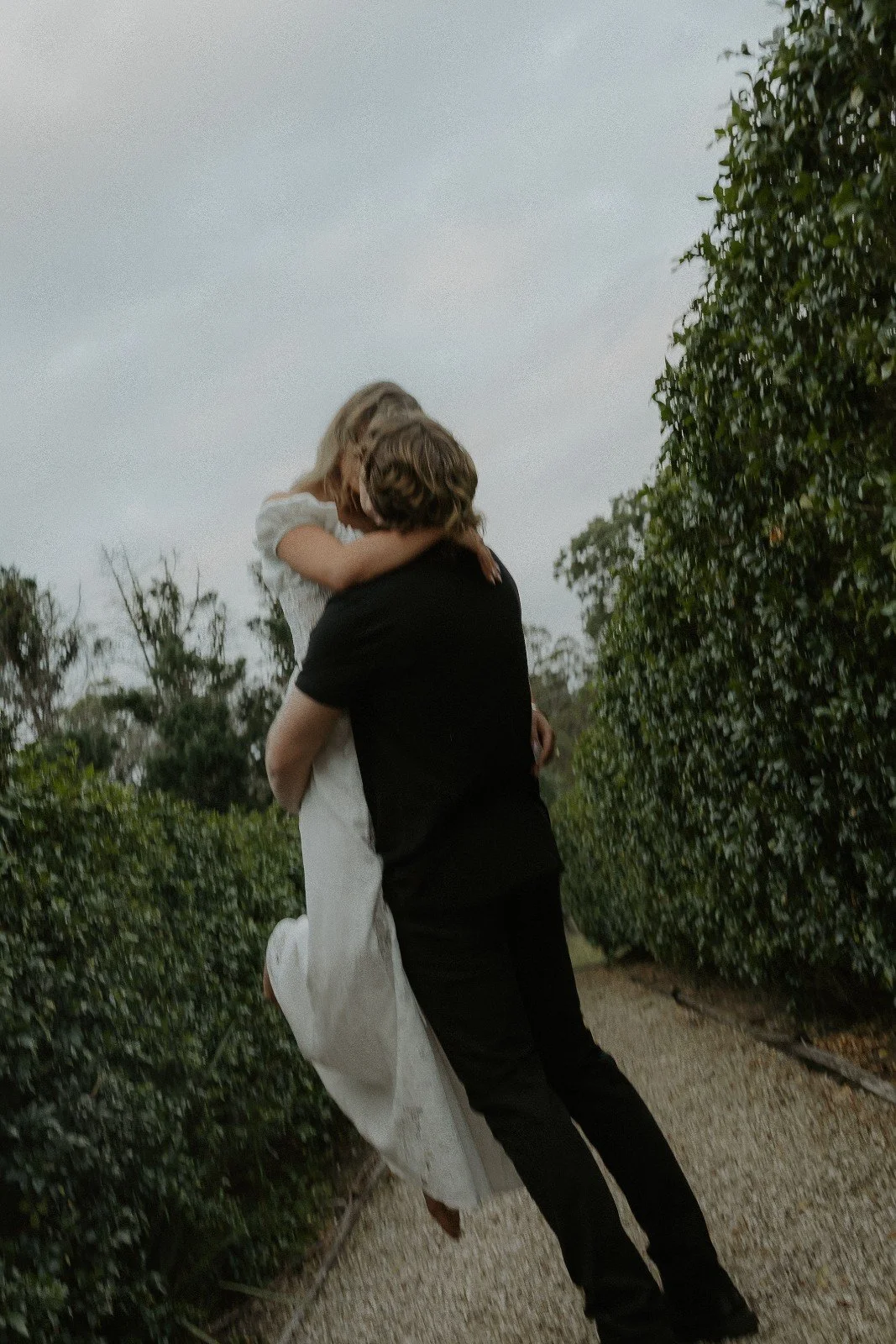 A person in black clothing holding and lifting a woman in a white dress in a forested outdoor setting, with trees and bushes around.