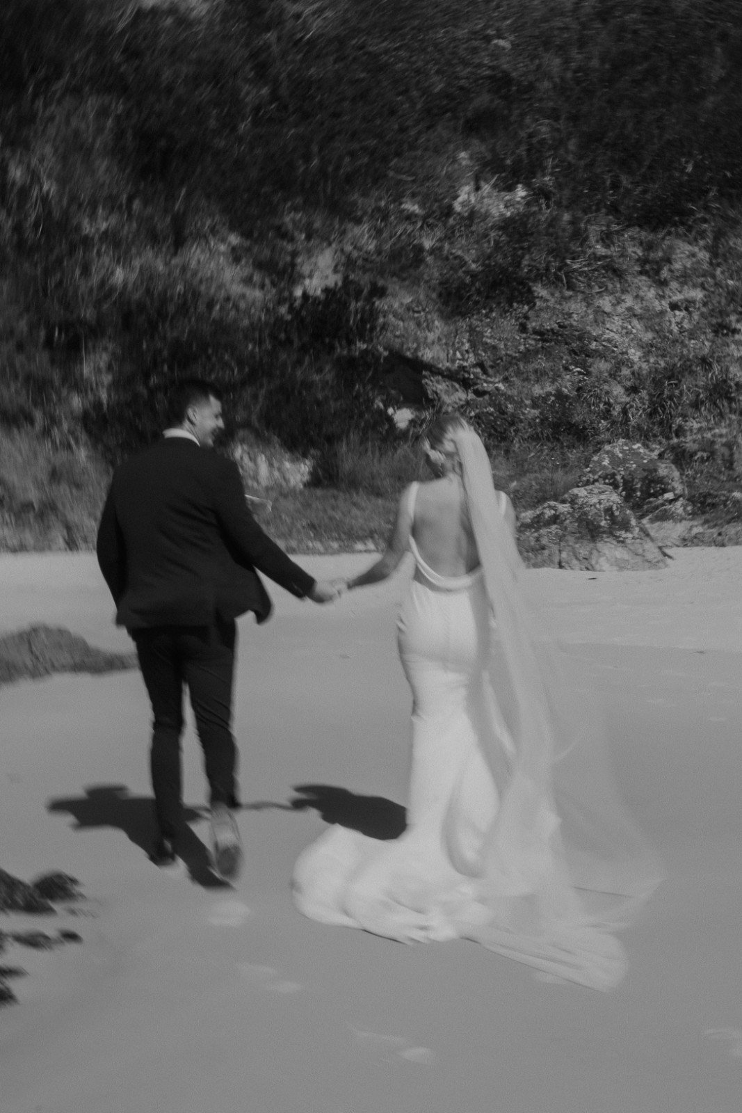 A black and white photo of a couple walking hand in hand on the beach, with the woman wearing a long white dress and the man in a dark suit. They are facing away from the camera, with trees and rocks in the background.