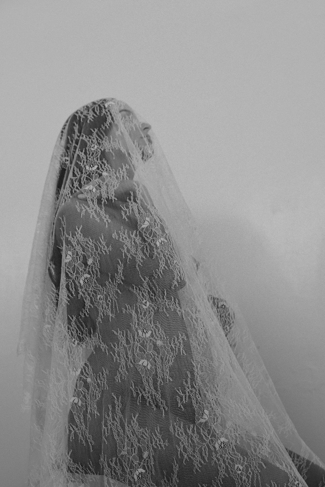 A woman in a wedding dress with a lace veil over her face, standing against a plain wall.