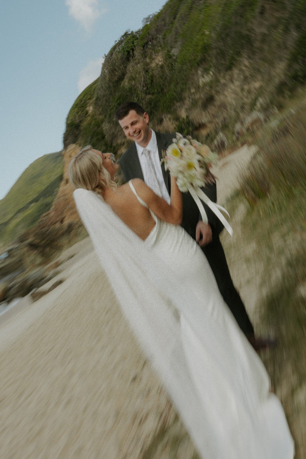 A bride and groom share a joyful moment on a beach, with the groom holding a bouquet and both looking at each other happily, scenic cliffs in the background.