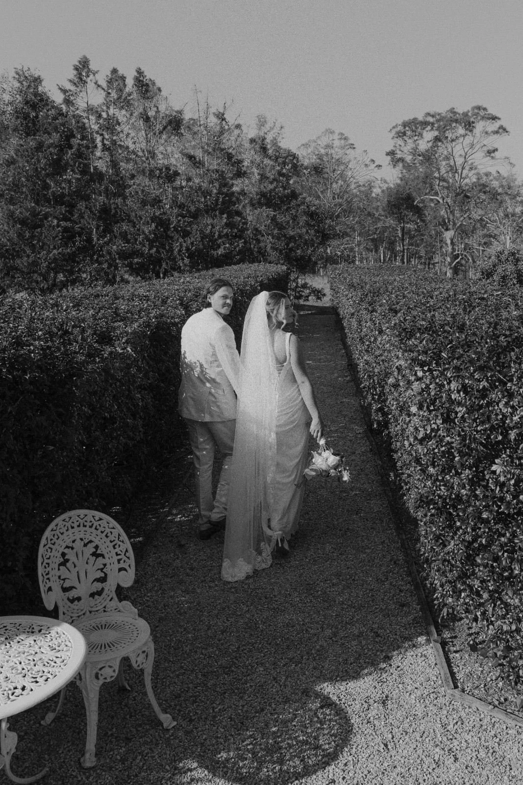 A bride and groom walking down a garden path, holding hands, with bushes and trees in the background. The bride is holding a bouquet, and there are decorative outdoor furniture pieces nearby.