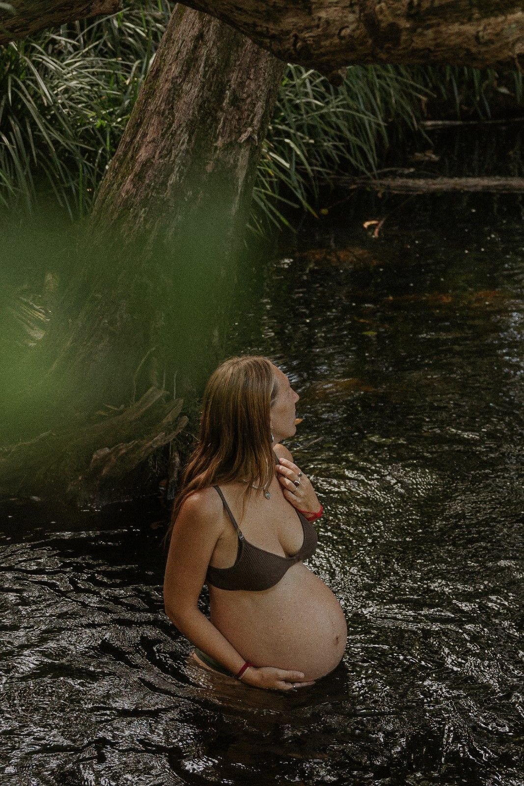 Pregnant woman standing in a river, holding her belly with one hand and touching her neck with the other in a natural outdoor setting.