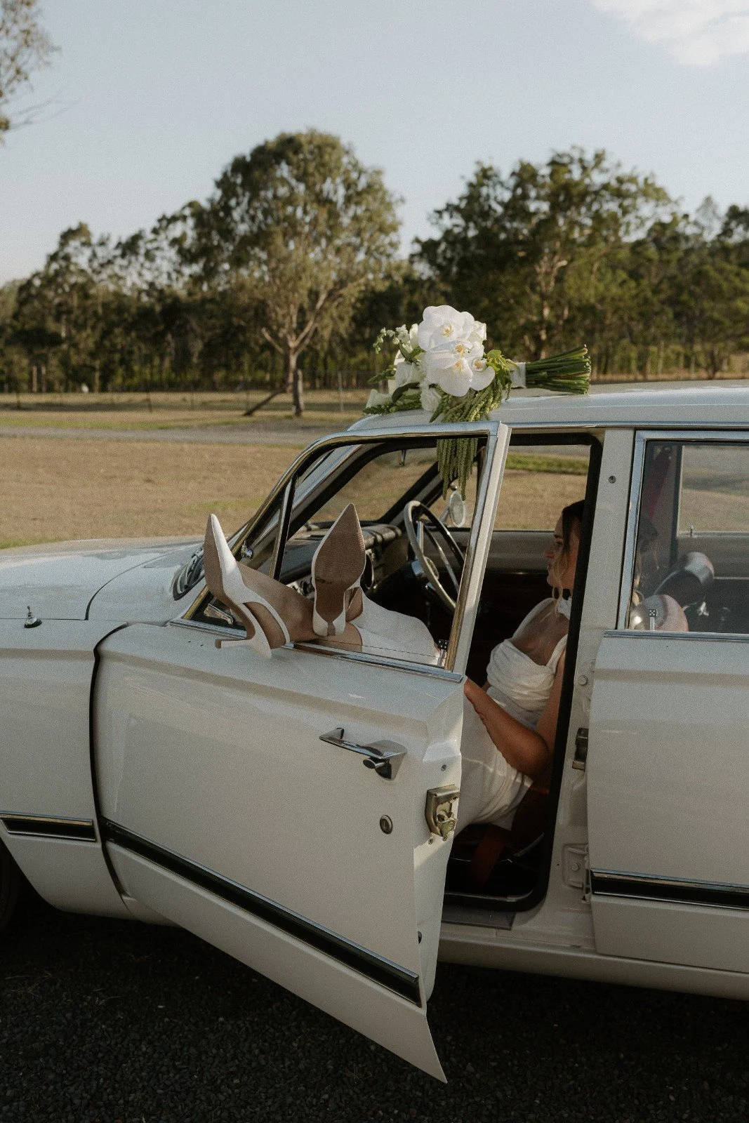 A woman in a white dress and high heels sitting in an open vintage white car with a bouquet of white flowers on the roof, in an outdoor setting with trees and grass.