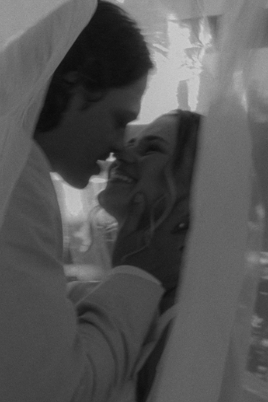 A black and white photo of a couple about to kiss, taken through a glass surface with reflections.