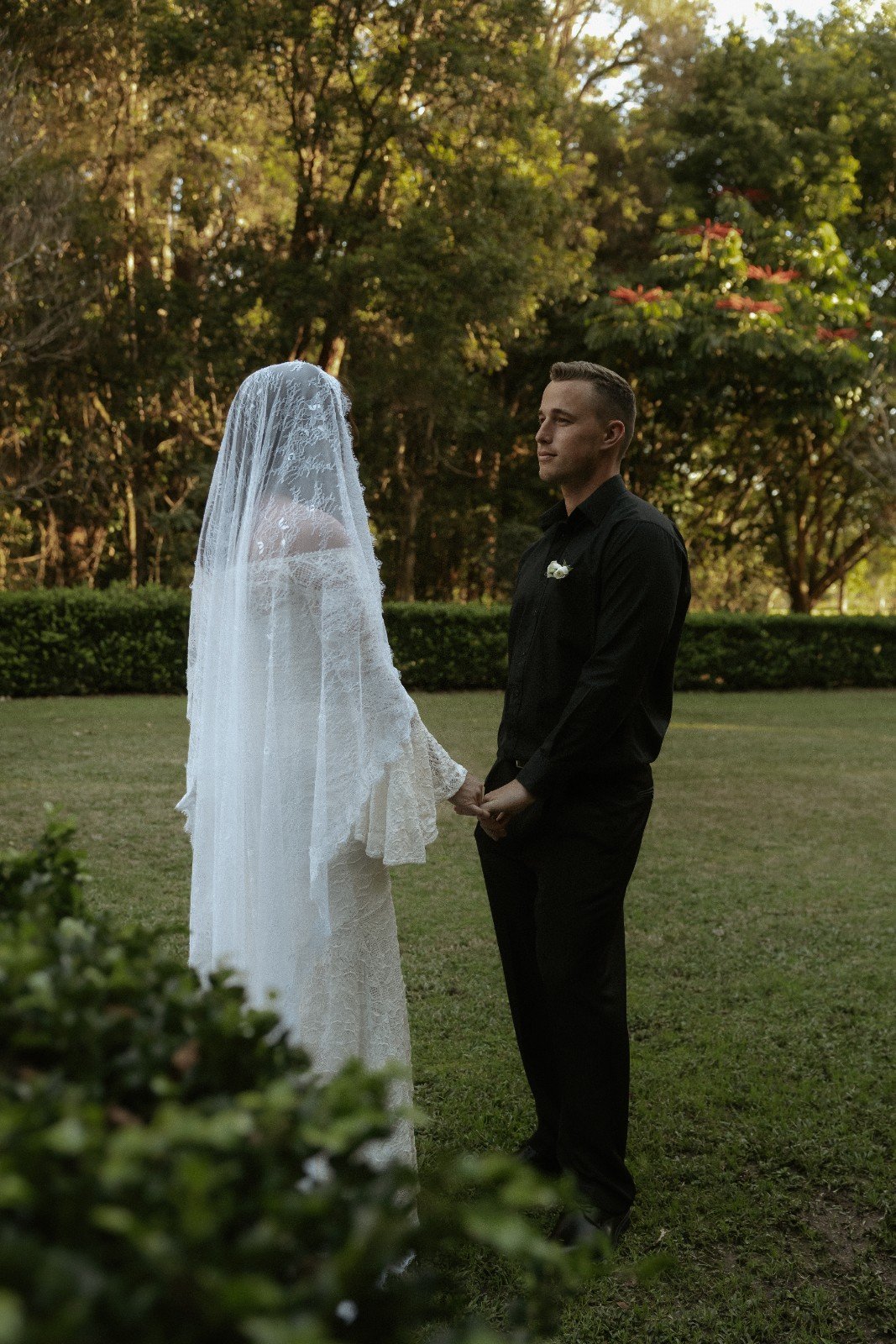 A bride and groom holding hands outdoors in a park during sunset, with trees and bushes in the background.