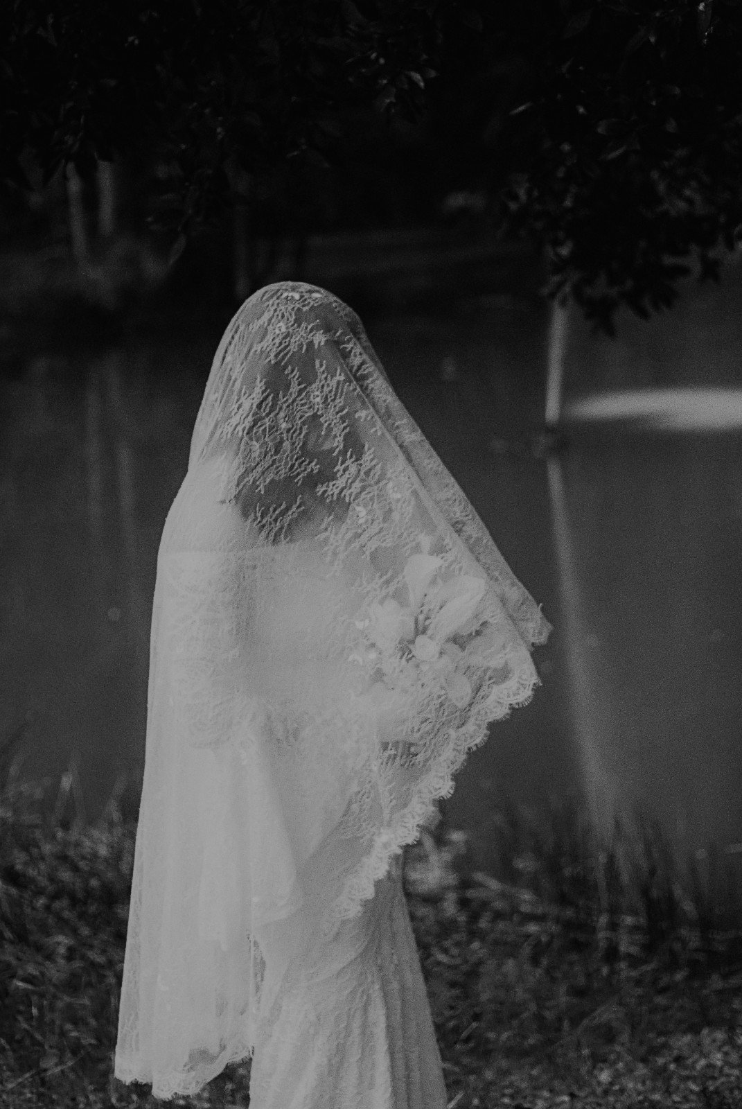 Black-and-white photo of a person in a lace wedding dress with a veil covering their head, standing outdoors at night near water.