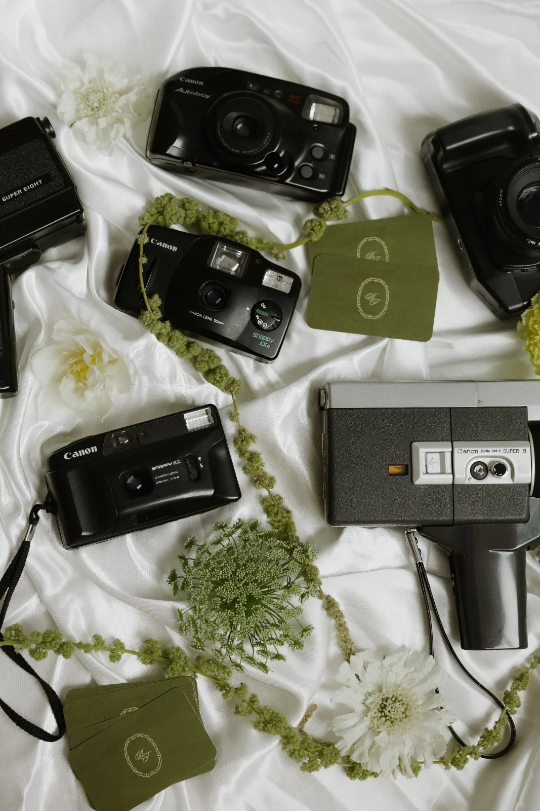 Multiple vintage cameras and instant cameras arranged on a white satin fabric, with white and green flowers and plant accents scattered around. Bloom Film Pohotgraphy collection.