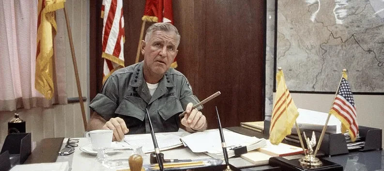 A man in a military uniform sitting at a desk with papers, flags, and office supplies in a room decorated with flags and a map on the wall.