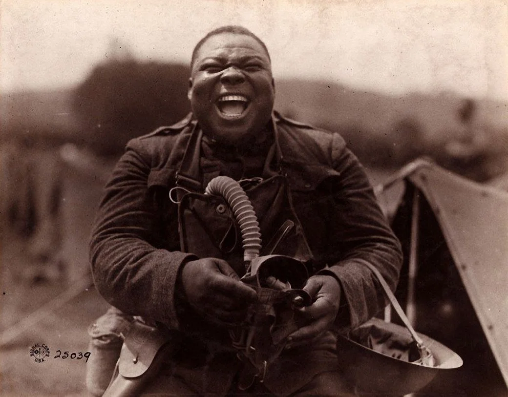 A man in military uniform smiling broadly while holding a mask or respirator, with a tent and soldiers in the background.