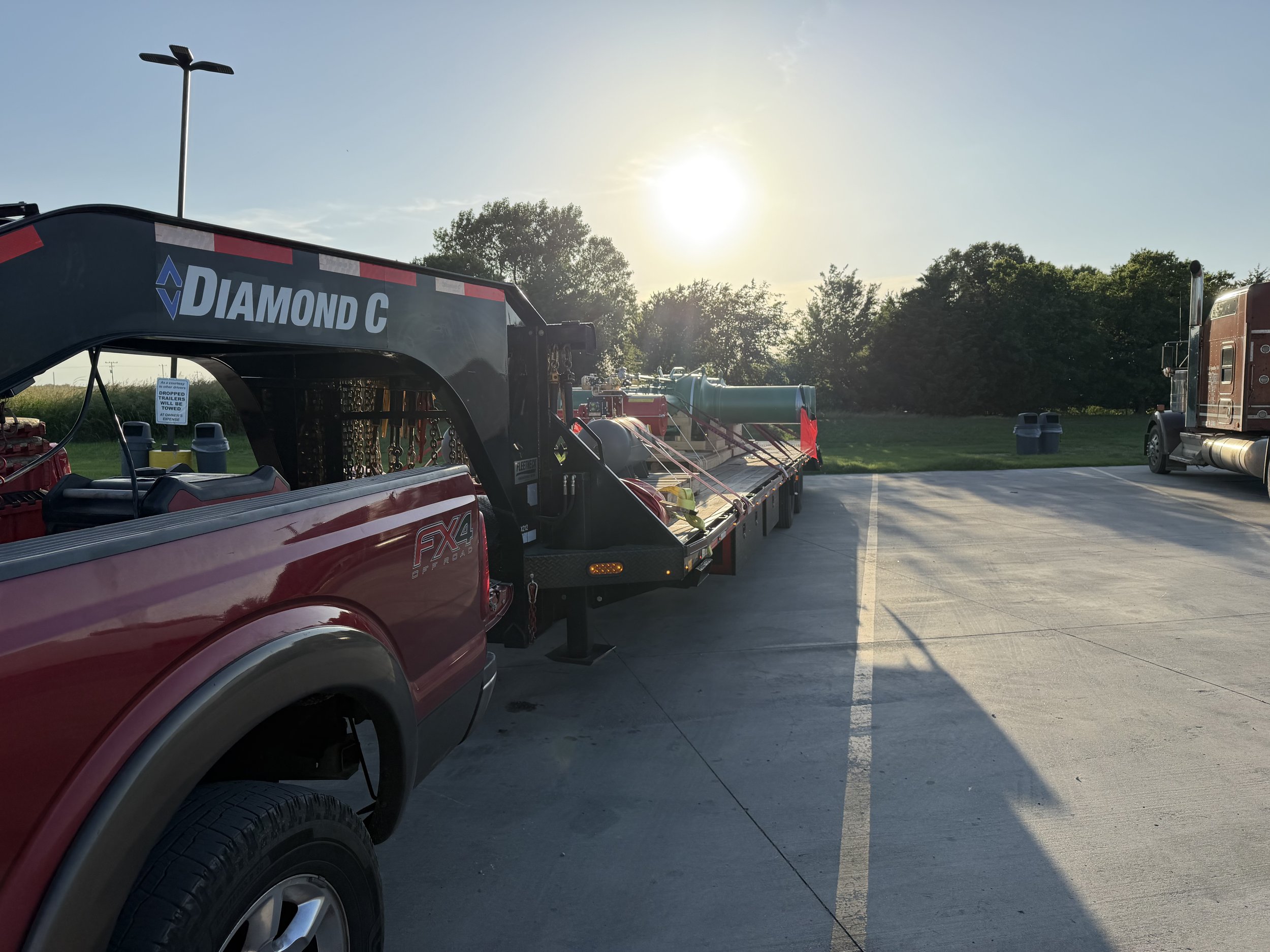 A red pickup truck with a flatbed trailer carrying equipment, parked in an empty parking lot during sunset with trees in the background.