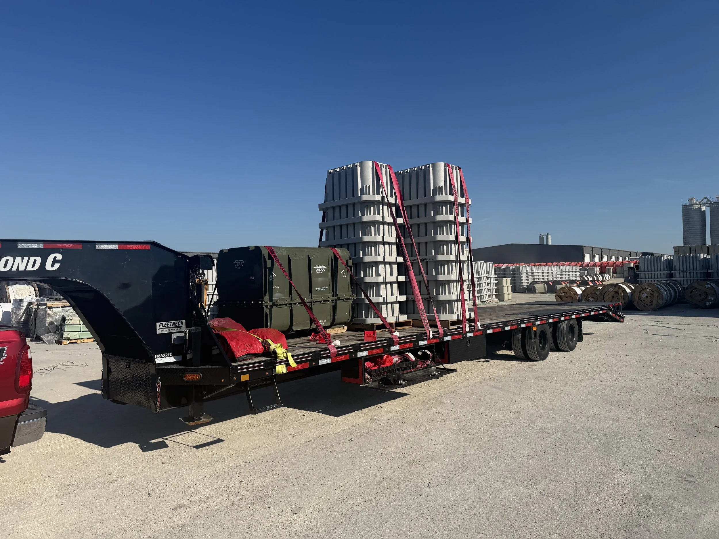 A flatbed truck carrying large plastic crates and storage containers, secured with pink straps, on a concrete surface under a clear blue sky with some industrial buildings in the background.