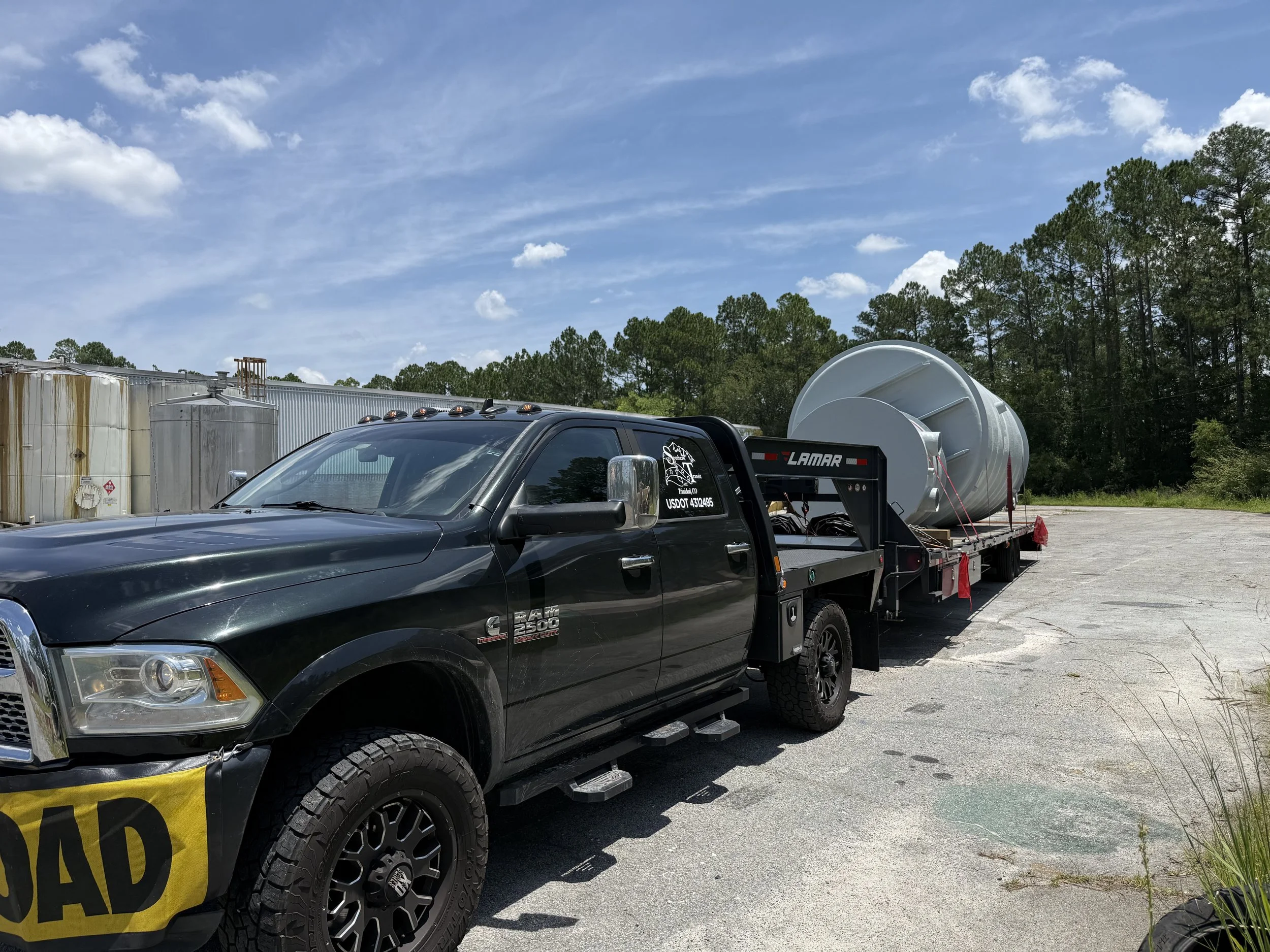 Black pickup truck with a large white tank on a flatbed trailer attached, parked outdoors near trees and a blue sky with scattered clouds.