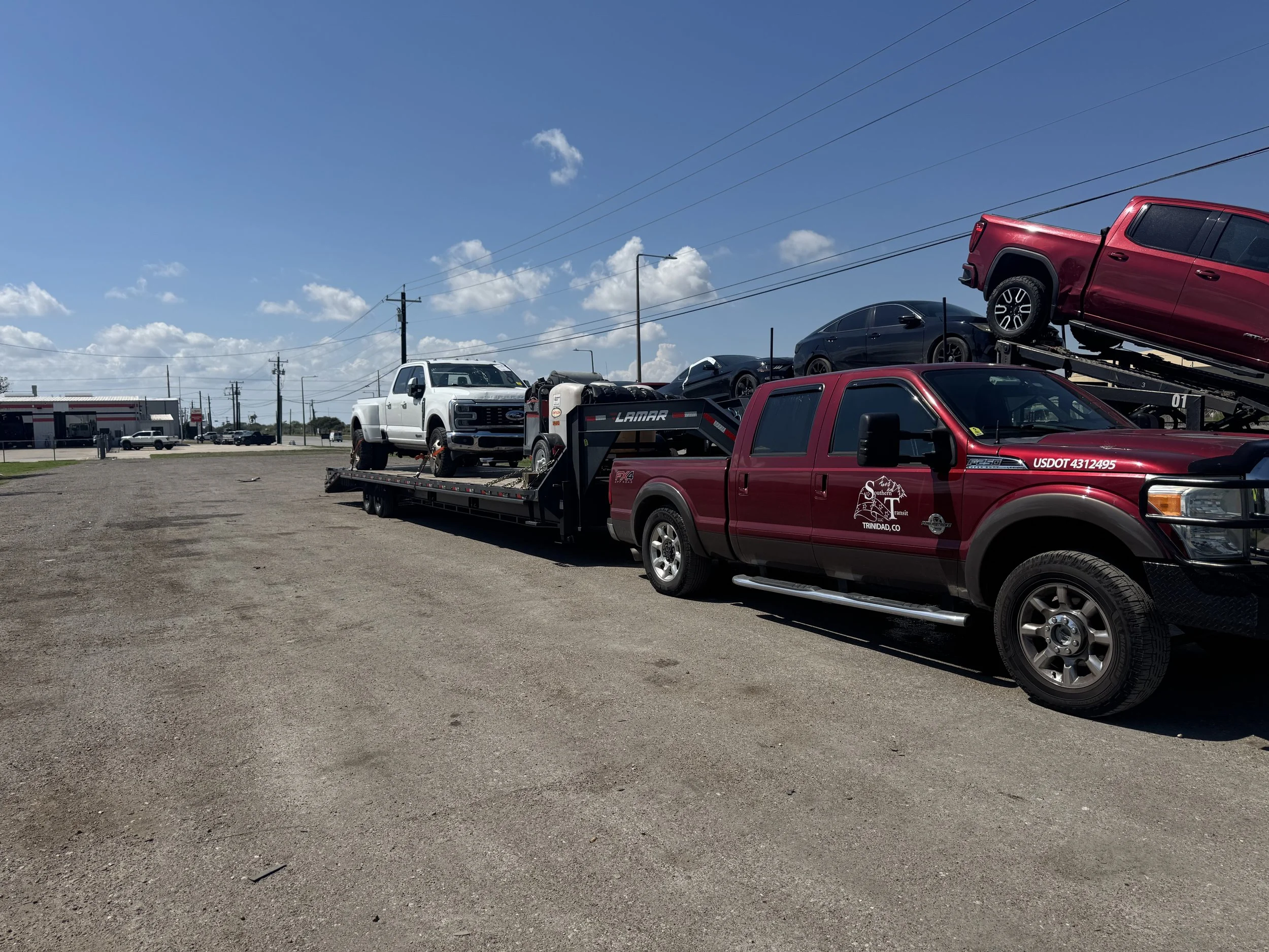 A red pickup truck transporting a black car and a white truck on a flatbed trailer in an open parking lot under a blue sky with scattered clouds.