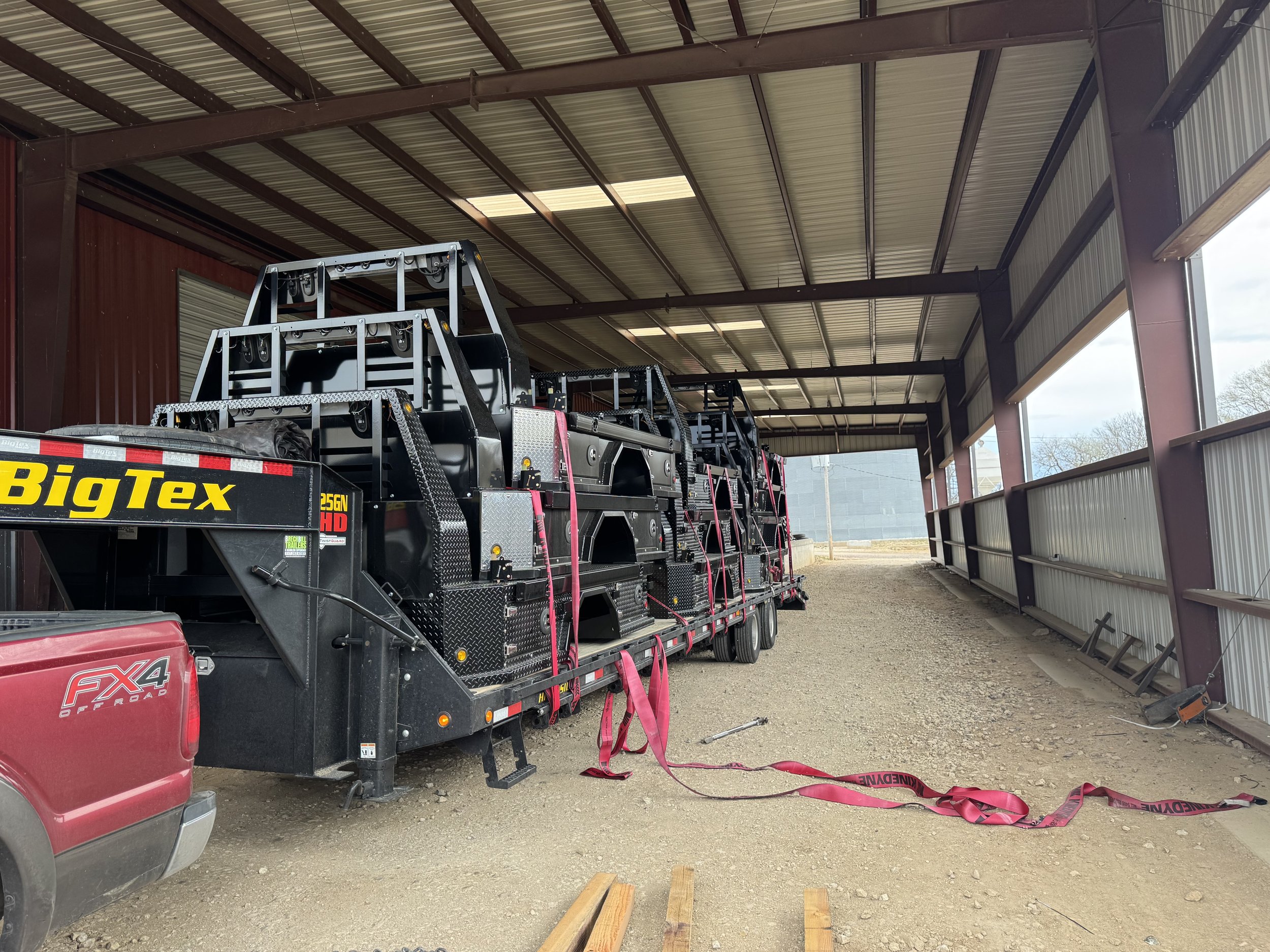 A flatbed trailer carrying multiple black construction lifts with the brand name Big Tex, parked inside a metallic building with a partially open side, red vehicle partially visible on the left, and wooden planks on the ground.