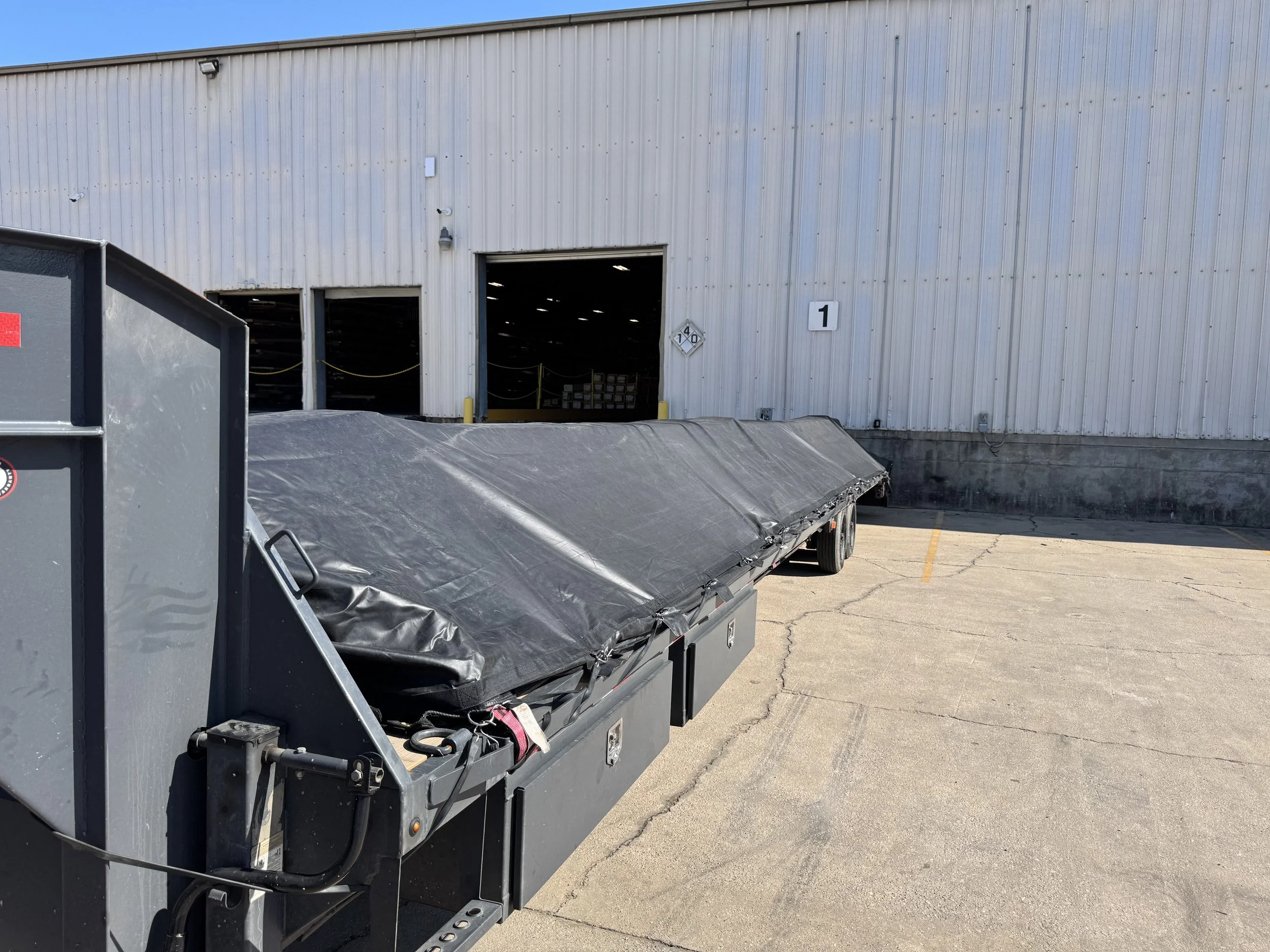 View of a large industrial warehouse with open door, items inside visible, and a semi-truck trailer parked outside on the concrete lot.