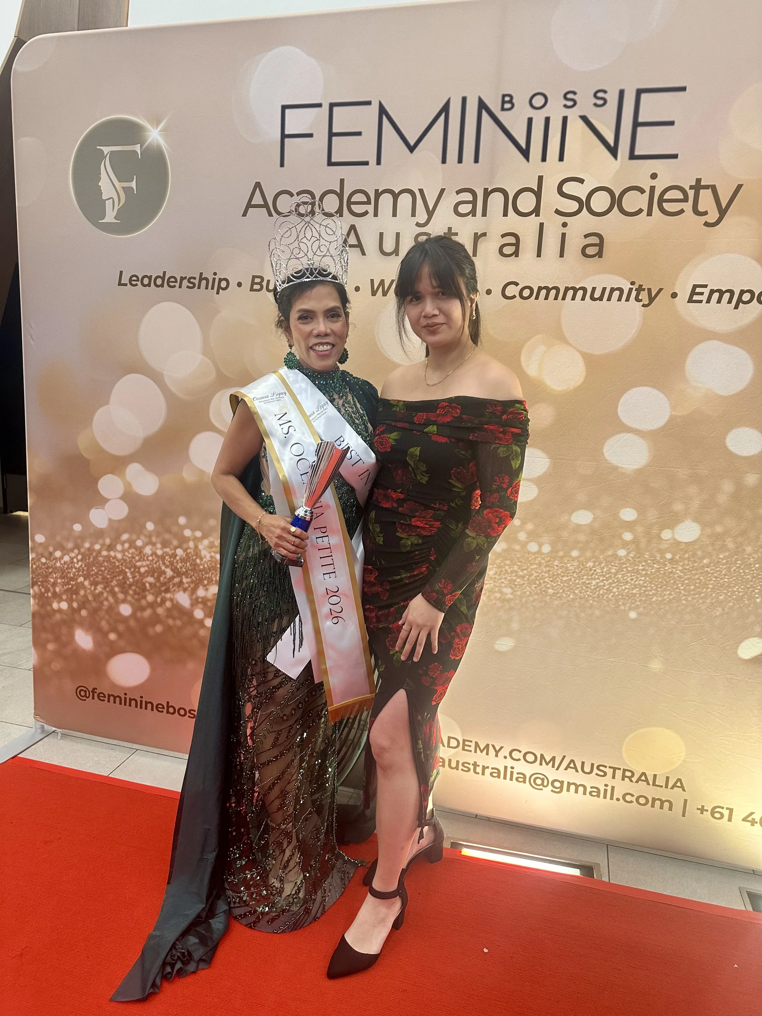 Two women standing together at an event, one wearing a crown and sash, and the other in a black off-the-shoulder dress with red floral pattern, both smiling for the photo in front of a backdrop for FemininE Boss Australia.