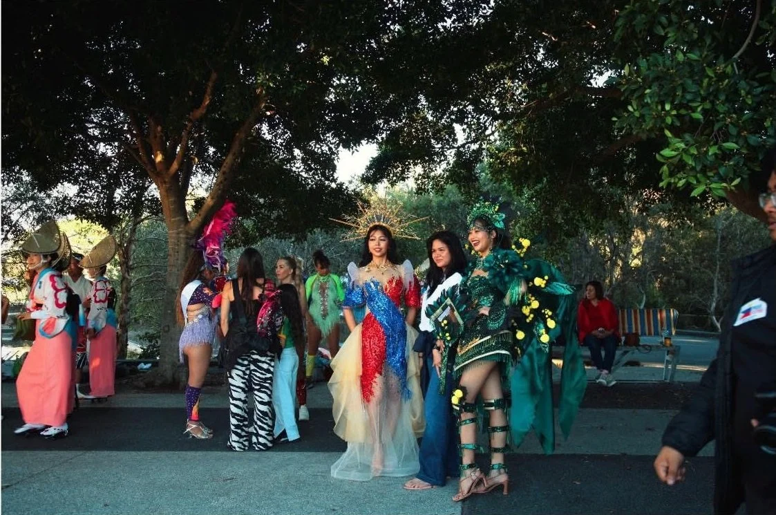 People dressed in colorful, elaborate costumes standing outdoors under trees, possibly at a cultural or fashion event.