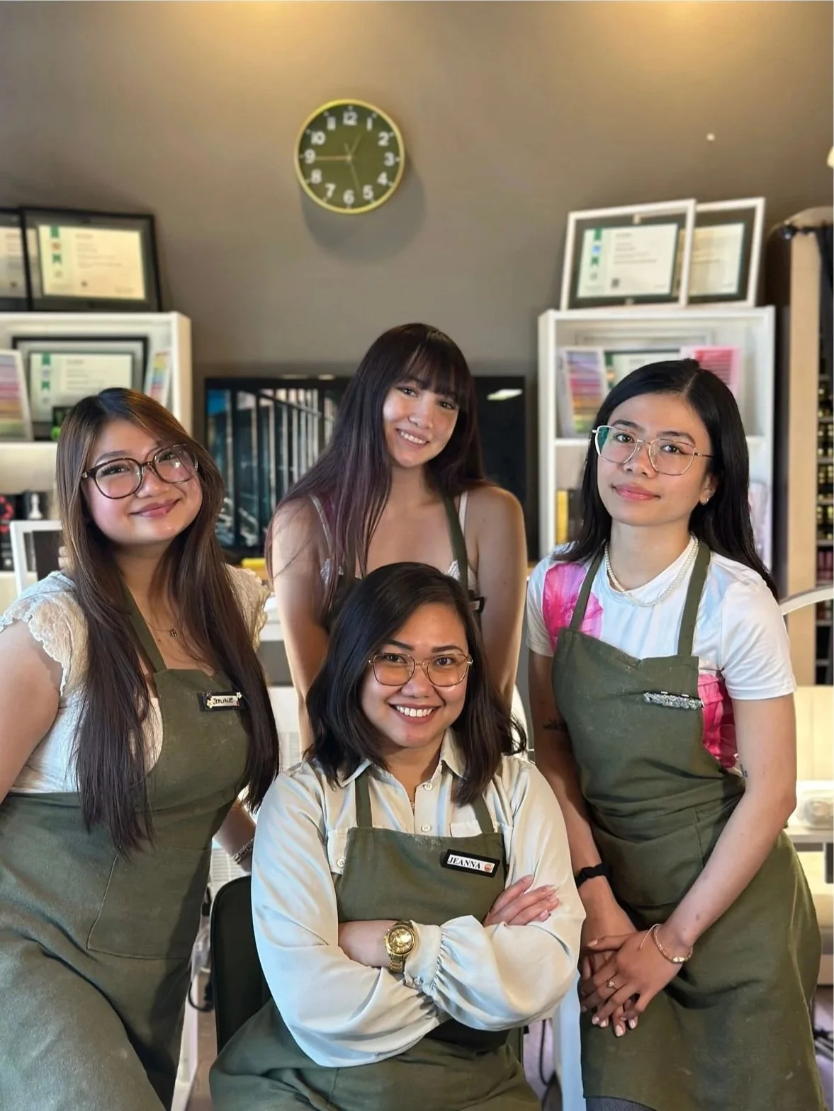 Four women smiling and posing for a photo inside a room, wearing green aprons, with a wall clock showing 1:55 behind them.