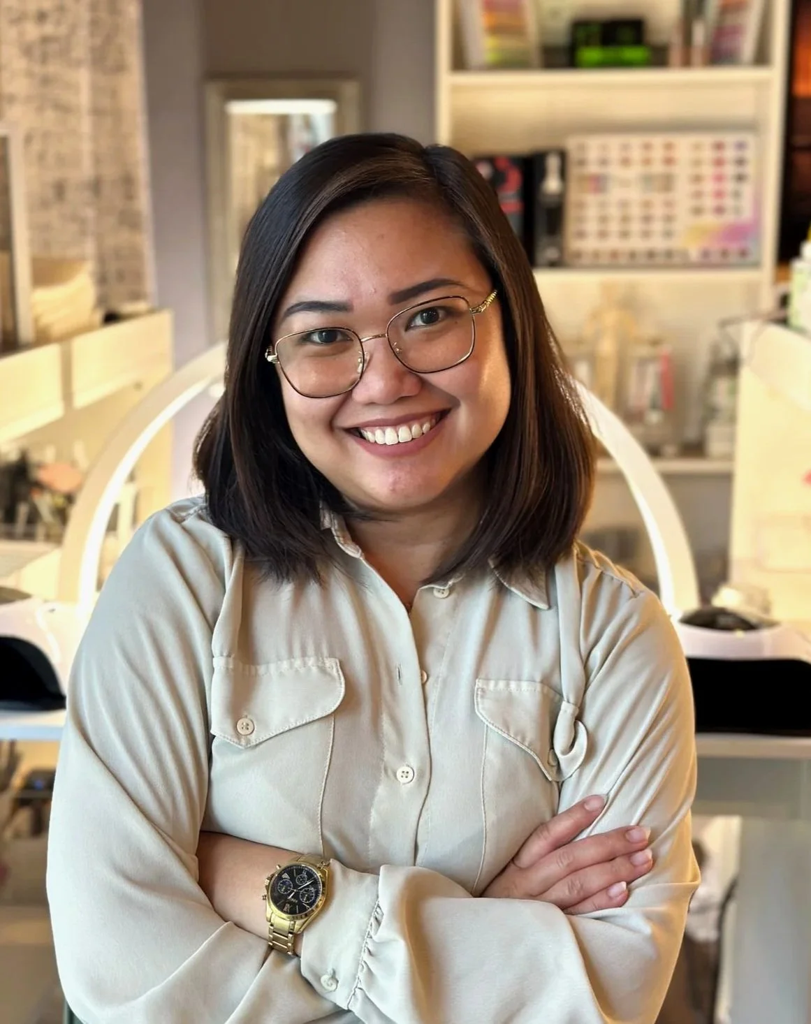 A woman with shoulder-length dark hair and glasses smiling with arms crossed, wearing a light-colored shirt, in a retail or office environment with shelves and products in the background.