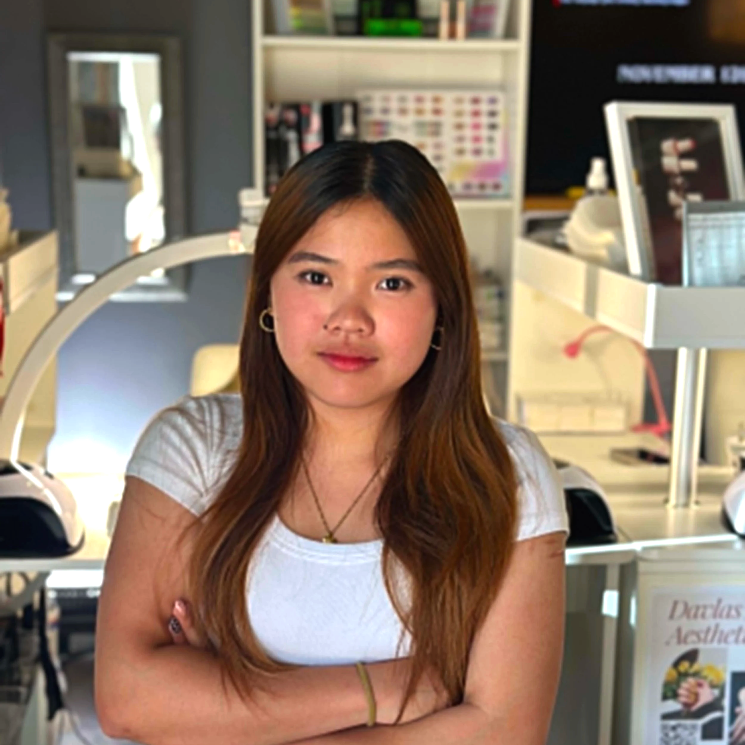 Front view of a woman with long brown hair wearing a white top, standing in a bookstore or library with bookshelves and framed photos or posters in the background.