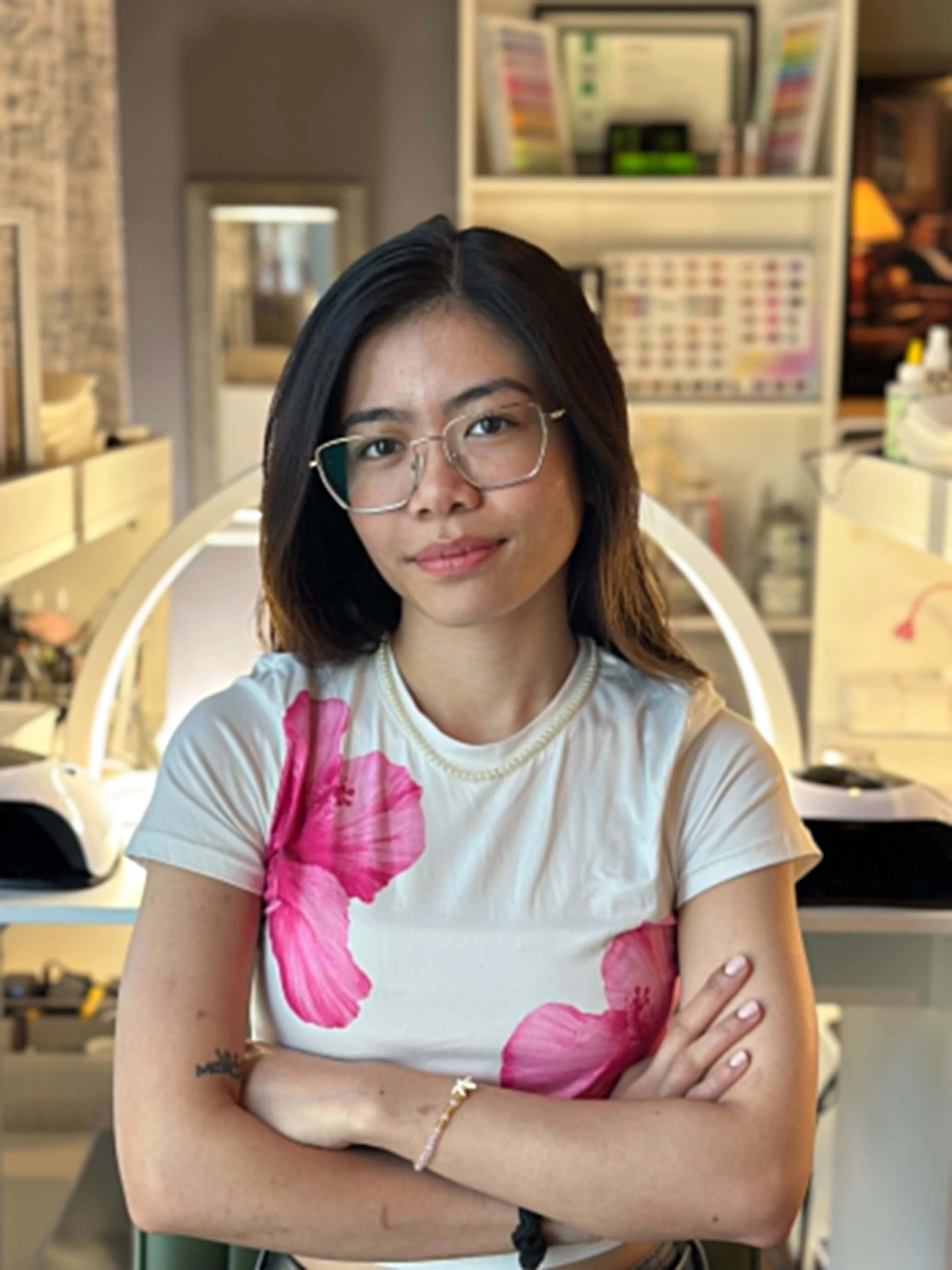 A young woman with black hair, glasses, and a floral shirt with pink flowers crosses her arms and stands in a room with shelves and various items in the background.