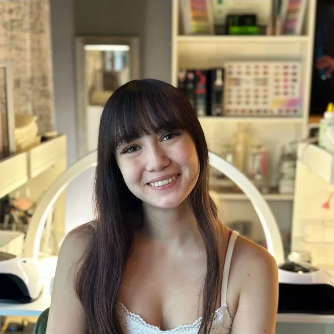 A young woman with long dark hair and bangs smiling at the camera, seated in a salon or beauty store with shelves of nail polish and beauty products in the background.