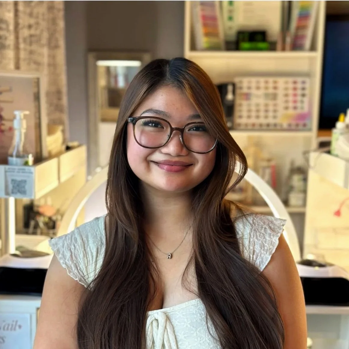 Smiling young woman with long brown hair and glasses, wearing a white top, in a cozy indoor setting with shelves and containers in the background.