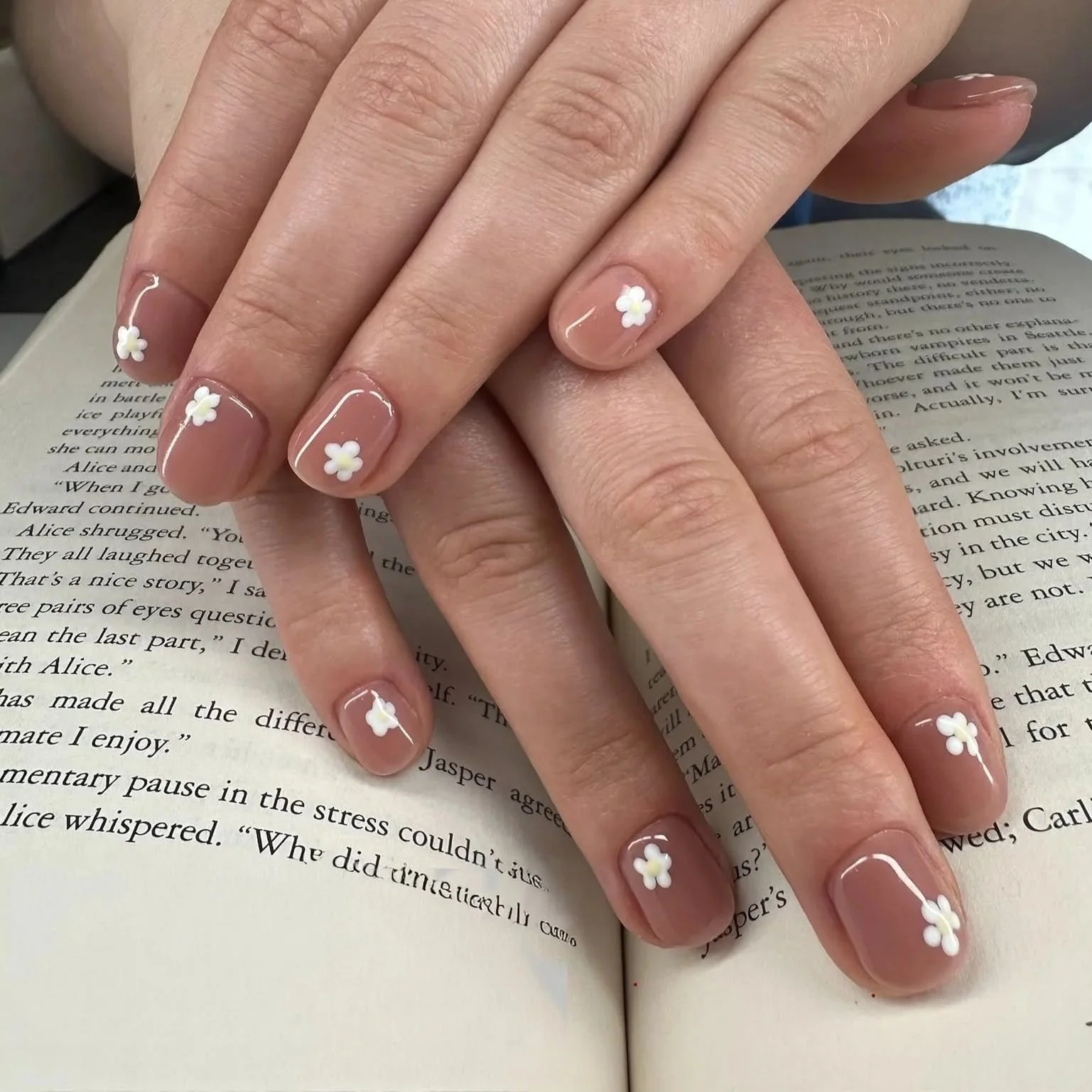 Hands with manicured nails resting on an open book, nails painted with clear polish and small white flower designs.