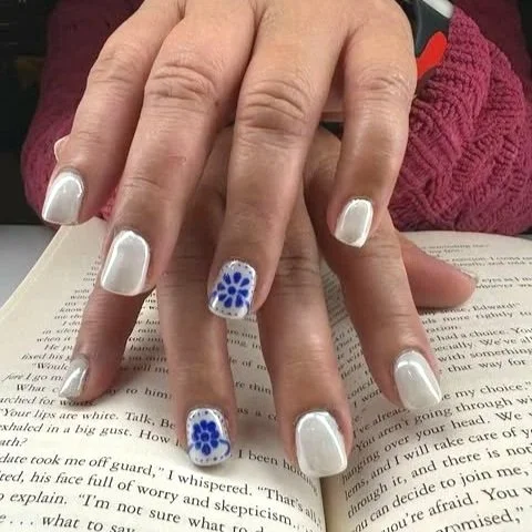 Close-up of hands with well-manicured nails, painted with white polish and a blue floral design on one nail, resting on an open book.