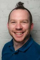 A smiling man with short hair and a beard, wearing a blue shirt, standing against a light-colored brick wall.