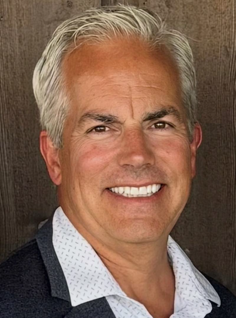 Close-up of a smiling middle-aged man with gray hair, wearing a white patterned shirt and dark blazer, against a wooden background.