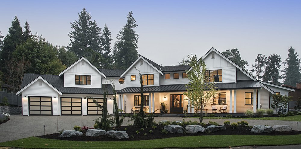 Modern two-story white house with black roof, three garage doors, front porch, and landscaped yard with rocks and trees.